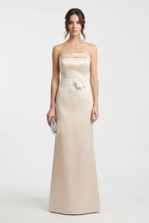 Vista frontal de modelo luciendo un vestido largo champán de fiesta con escote palabra de honor y flor decorativa en el cinturón.