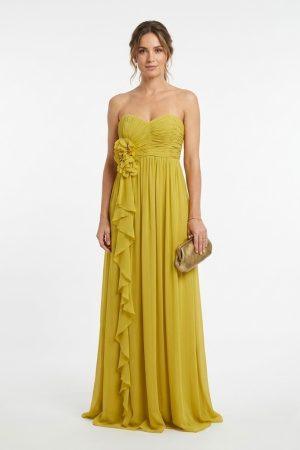 Mujer luciendo un elegante vestido largo de fiesta amarillo mostaza con escote palabra de honor y detalle de flor de frente.