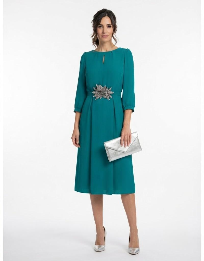 Modelo vistiendo un diseño midi en color verde esmeralda con un aplique floral decorativo en la cintura y bolso plateado.